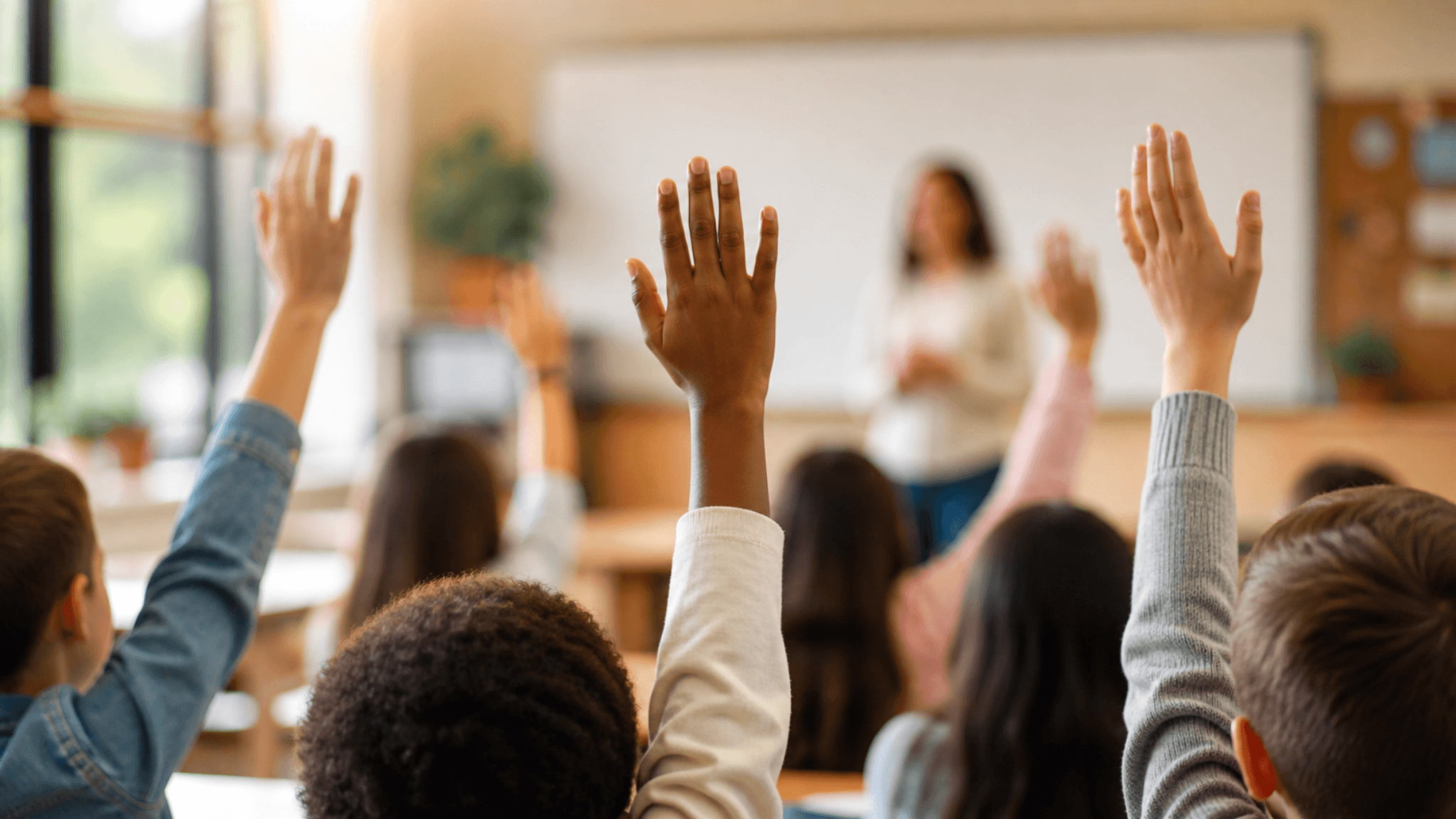 Estudiantes levantando la mano en un aula mientras una maestra está al frente